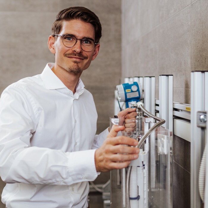 A man in a white shirt holding a hose and smiling, standing in front of control panels with buttons.
