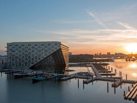 Modern geometric building beside a calm waterway during sunset, with docks and boats in the foreground.