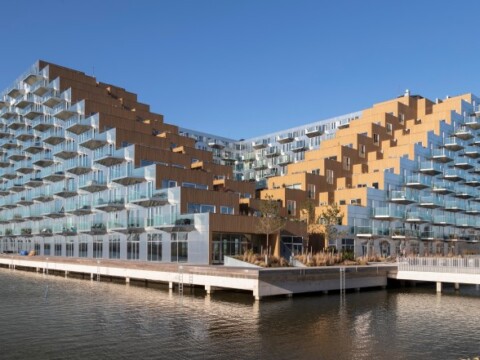Modern architectural building with angled terraces and large glass windows, reflecting over water under a clear blue sky.
