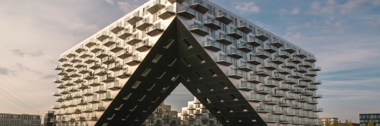 Modern architectural building with a distinctive triangular base, featuring numerous balconies and large glass windows, situated near water.