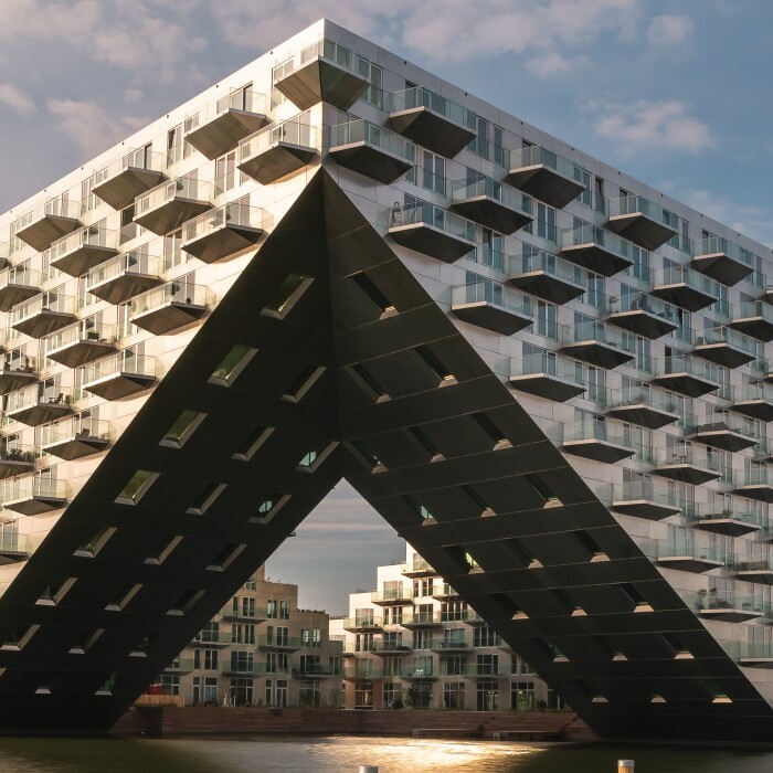 Modern architectural building with a distinctive triangular base, featuring numerous balconies and large glass windows, situated near water.