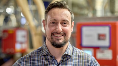 A man with a beard and short hair smiles while standing in an industrial setting with equipment in the background.