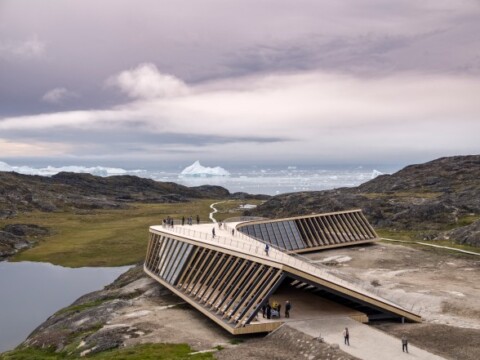 A modern architectural structure with large angled roofs, set in a rocky landscape with green grass and a lake, surrounded by icebergs in the distance under a cloudy sky.
