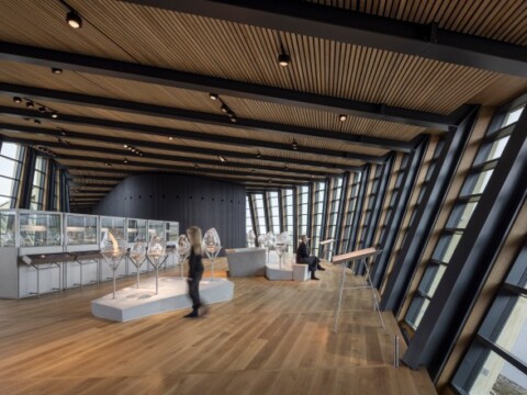 A modern exhibition space with large windows showcasing artworks and installations, featuring wooden flooring and minimalistic design.