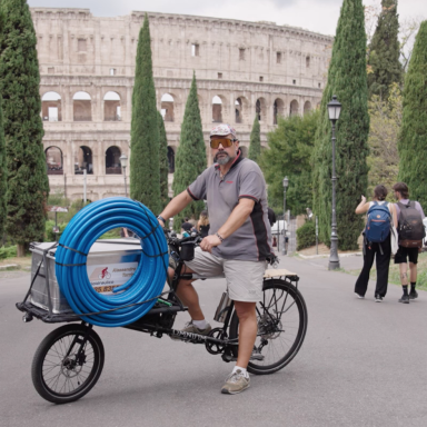 Ein Mann auf einem Fahrrad mit blauen Schläuchen, im Hintergrund das Kolosseum und Bäume.