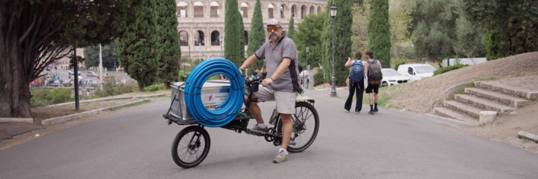 Ein Mann fährt auf einem Lastenrad mit blauen Rohren durch eine Strasse, wobei das Kolosseum im Hintergrund sichtbar ist.