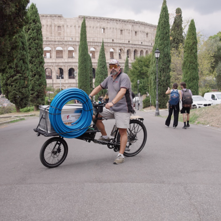 Ein Mann fährt auf einem Lastenrad mit blauen Rohren durch eine Strasse, wobei das Kolosseum im Hintergrund sichtbar ist.