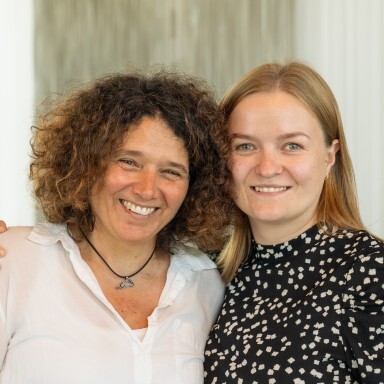 Two women standing closely together, smiling warmly at the camera, with one wearing a white shirt and the other a black with a white polka dot pattern.