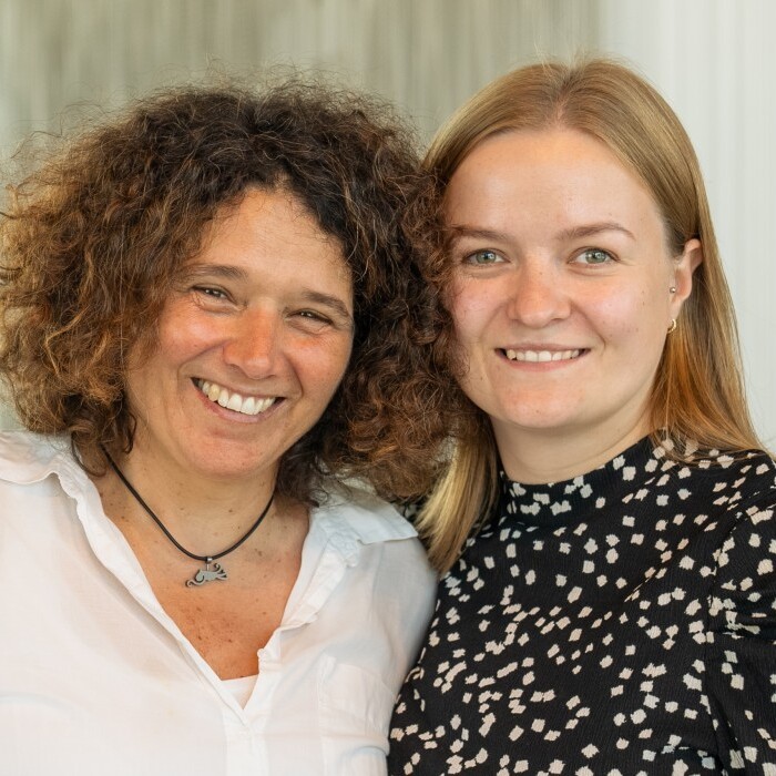 Two smiling women pose together, one with curly hair and a white shirt, the other with straight blonde hair and a black polka dot top, against a bright indoor backdrop.