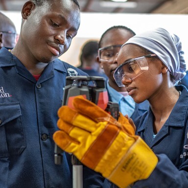 A group of individuals in blue work uniforms, with one person wearing goggles and gloves, examines a tool in a workshop setting.