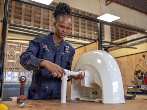 A woman in a blue work jacket assembling a plumbing fixture in a workshop.