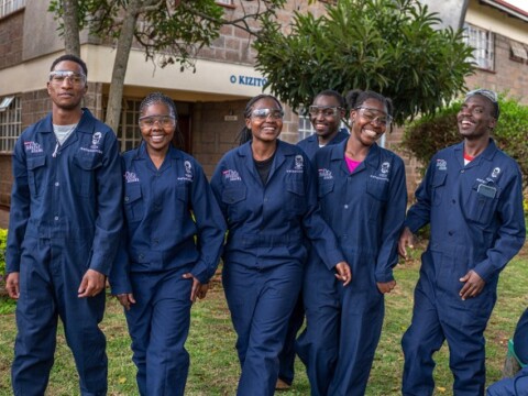 A group of people in blue overalls smiling and walking together outdoors near a building and greenery.