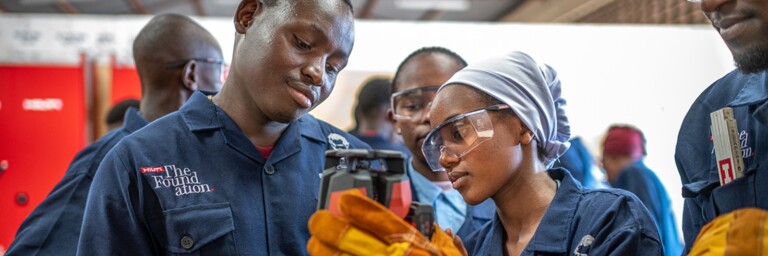 A group of young adults in blue work uniforms and safety goggles, examines a tool.