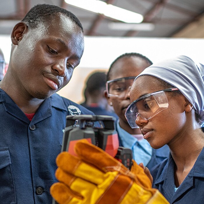 A group of young adults in blue work uniforms and safety goggles, examines a tool.