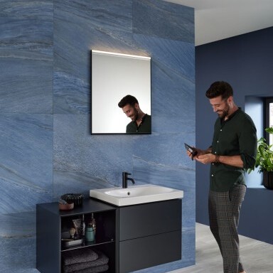 Ein Mann steht barfuss in einem modern gestalteten Badezimmer vor einem Waschbecken und schaut auf sein Handy.