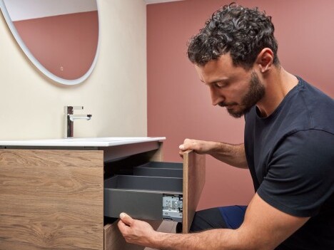 Ein Mann mit lockigem Haar, der an einem modernen Holzschrank mit geöffneten Schubladen in einem Badezimmer arbeitet.