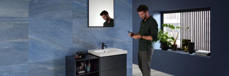 Ein Mann steht barfuss in einem modernen Badezimmer, schaut auf sein Smartphone und lächelt, während er vor einem Waschbecken mit schwarzem Unterschrank steht.