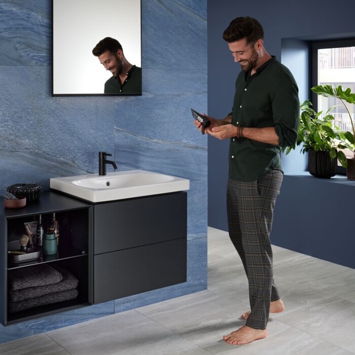 Ein Mann steht barfuss in einem modernen Badezimmer, schaut auf sein Smartphone und lächelt, während er vor einem Waschbecken mit schwarzem Unterschrank steht.