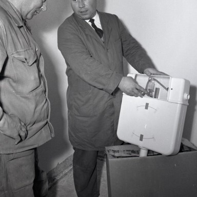 Two men are discussing a product, with one man holding the product and demonstrating something to the other, in a plain room.