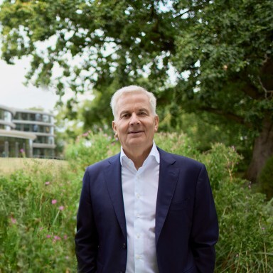 A middle-aged man in a dark suit stands confidently amidst greenery, with a modern building visible in the background.
