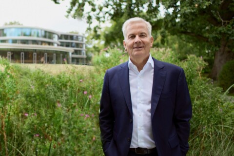 A man in a suit stands in front of a modern building surrounded by greenery.
