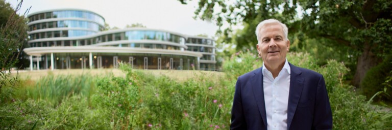 A man in a suit stands in front of a modern building surrounded by greenery.