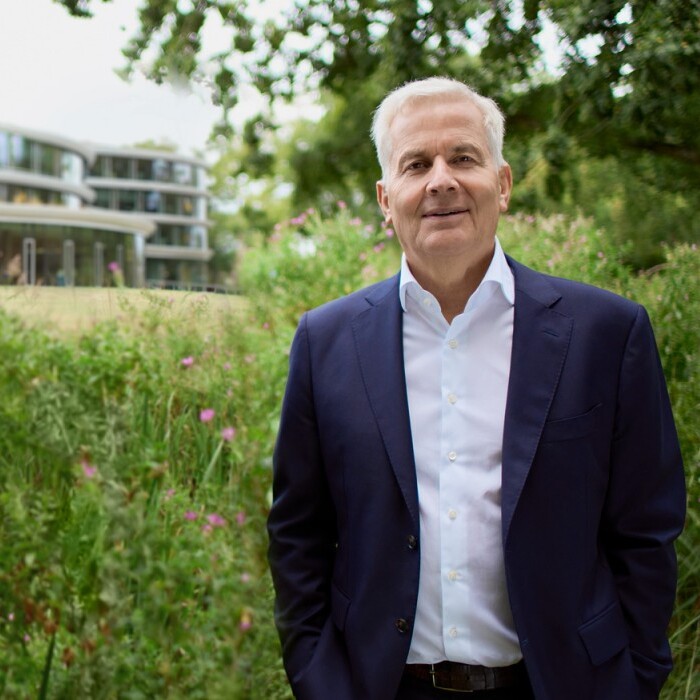 A man in a suit stands in front of a modern building surrounded by greenery.