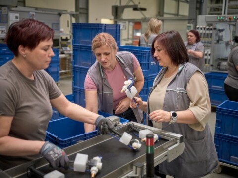 Three women in work attire collaborating on an assembly line with equipment and blue storage bins in a factory setting.
