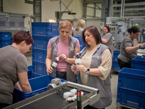 Women working in a factory setting, inspecting items and interacting around a conveyor belt, with blue storage containers in the background.