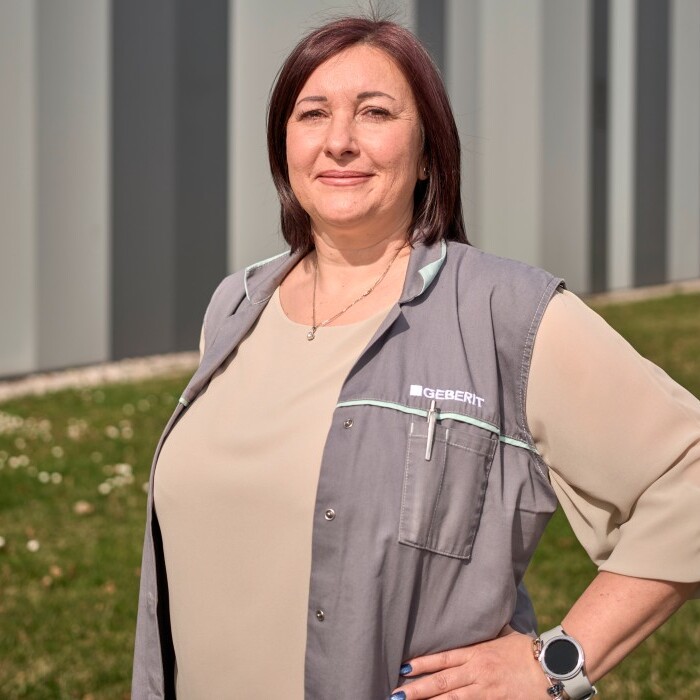 A woman in a gray vest stands confidently outdoors, with a grassy area and a modern building in the background.