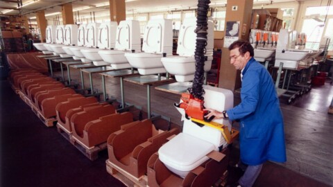 A factory setting with a worker in a blue coat operating machinery to package toilets, with rows of toilets in the background.