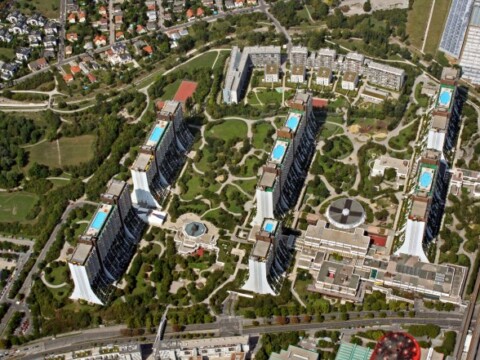 Aerial view of several high-rise buildings arranged around green spaces and swimming pools, with a neighborhood in the background.