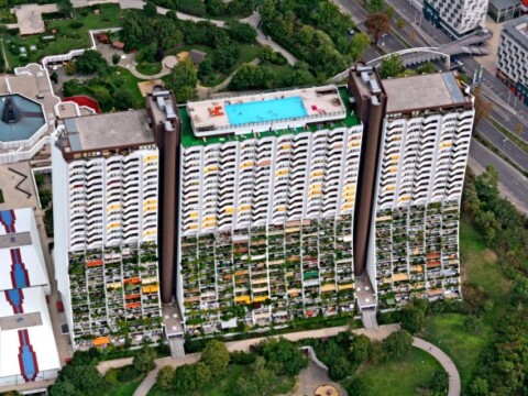 Aerial view of a modern building with balconies featuring lush green plants, and a rooftop pool surrounded by colorful sun loungers.