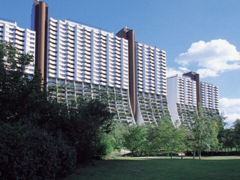 Modern multi-story residential buildings viewed from a grassy area, with trees and a blue sky.