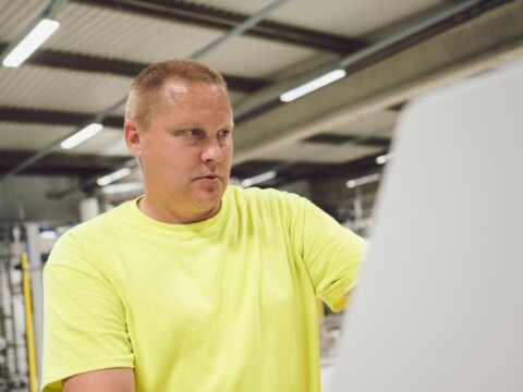 Ein Mann in einem hellgelben T-Shirt arbeitet konzentriert an einer Maschine in einer Produktionshalle.