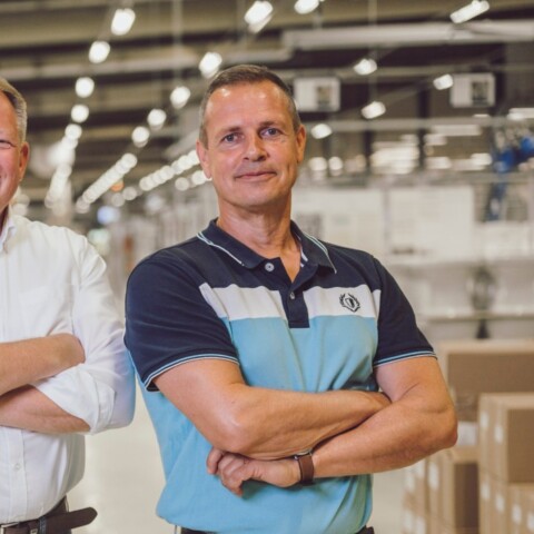 Drei Männer stehen in einem modernen, hellen Innenraum einer Fabrik oder Lagerhalle, die Arme verschränkt und lächeln.