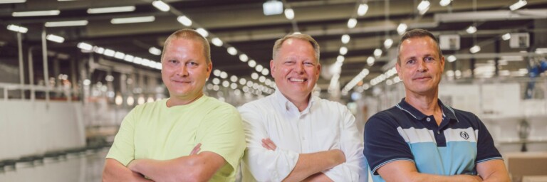 Drei Männer stehen in einem modernen, hellen Innenraum einer Fabrik oder Lagerhalle, die Arme verschränkt und lächeln.