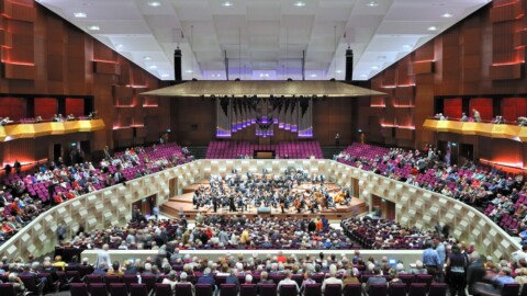 A large concert hall filled with an audience watching an orchestra perform on stage.