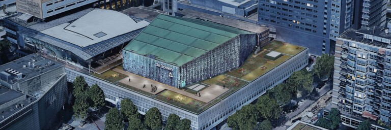 Aerial view of a large modern building with a green roof, surrounded by urban architecture and trees.
