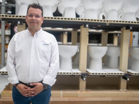 A man in a white shirt stands with hands clasped in front of shelves displaying white toilets.