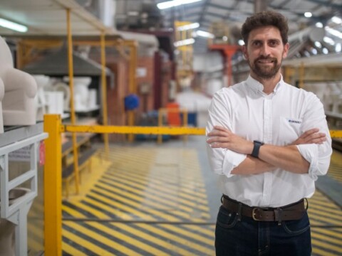 A man with arms crossed stands confidently in a factory setting, surrounded by industrial machinery and safety markings on the floor.