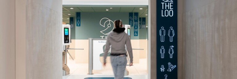 A person walks towards a restroom entrance, with directional signage on the walls indicating bathroom options and accessibility features.