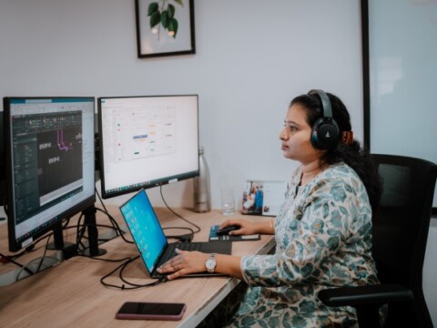 A woman wearing headphones works at a desk with two computer monitors.