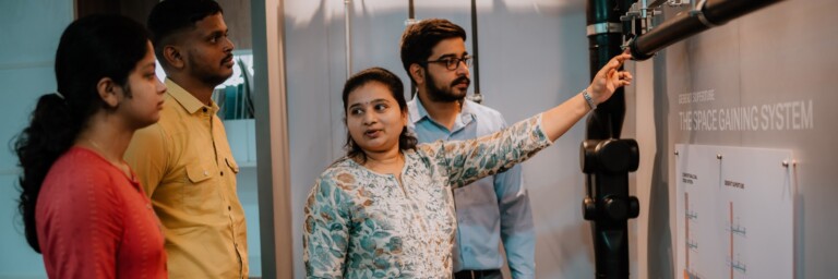 A group of four individuals stands around a display, with one person pointing to a pipe system.