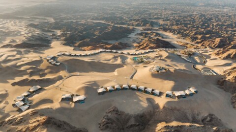 Aerial view of a desert landscape featuring a series of luxury tents arranged in a winding formation amidst rocky terrain and sand dunes.