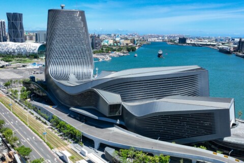 A modern architectural building with a unique, curved design situated near water, surrounded by city skyline and greenery.