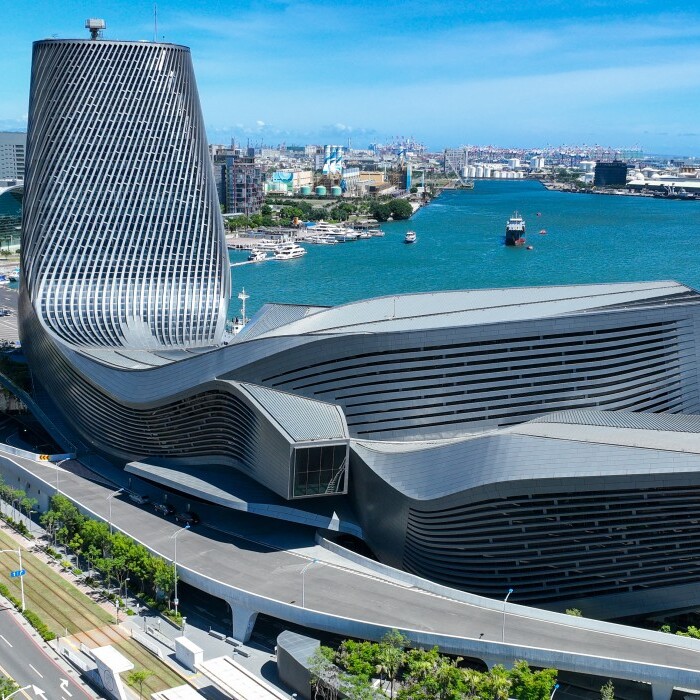 A modern architectural building with a unique, curved design situated near water, surrounded by city skyline and greenery.