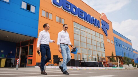 Two men in white shirts and jeans walking in front of a brightly colored store facade.
