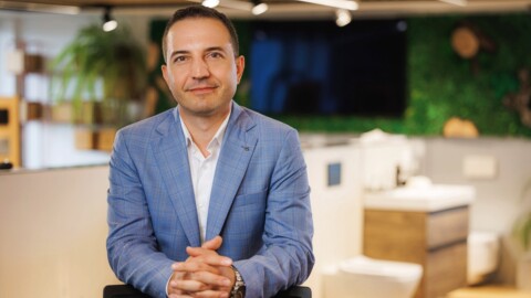 A man in a light blue suit is sitting with his hands clasped, smiling in a modern office environment with green plants in the background.
