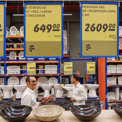 Ein Verkaufstisch mit mehreren grossen Schalen und zwei Mitarbeitern in einem Geschäft, Hintergrund mit Preisschildern für Sanitärprodukte.